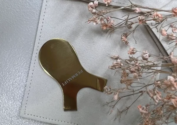 Close-up of a gold Permalift metal Y-comb resting on a light gray surface, surrounded by dried flowers, designed for precise lash lift treatments