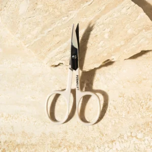 Permalift scissors with a sleek design resting on a textured beige surface, ideal for precise brow shaping and trimming