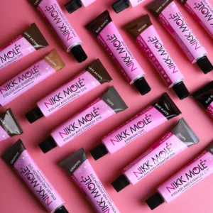 Collection of Nikk Mole Eyelash and Eyebrow Dye tubes in vibrant pink packaging, featuring a variety of shades including dark brown, graphite, blue black, and light brown on a pink background