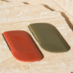 Permalift Duo Brow Razors in earthy terracotta red and olive green shades, displayed on a textured beige stone surface, showcasing their sleek and ergonomic design